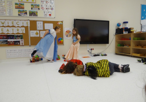 Laura w stroju pszczoły i Oliwia w stroju biedronki leżą na ziemi. Zuzia i Matylda odgrywają scenkę.